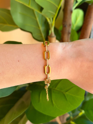 A gold chain bracelet with elongated rectangular links and a small dangling charm, shown worn on a person's wrist in front of large green leaves.