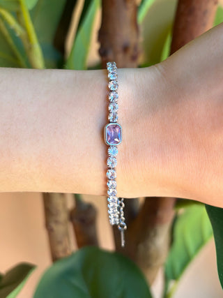 A delicate silver beaded bracelet with a central lavender-purple oval bead, worn on a person's wrist against a background of green leaves and brown tree branches.