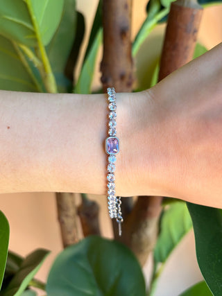 A delicate silver beaded bracelet with a central lavender-purple oval bead, worn on a person's wrist against a background of green leaves and brown tree branches.