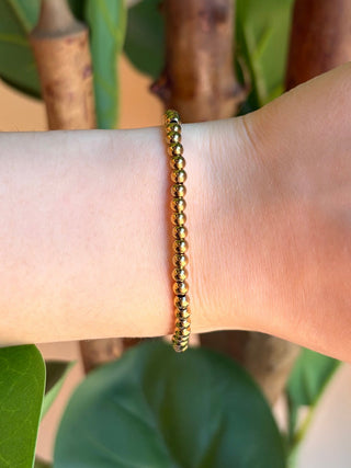 A delicate gold beaded chain bracelet worn on a person's wrist, shown against a background of green leaves and brown tree trunks.