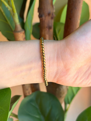 A gold beaded chain bracelet worn on a person's wrist, shown against a background of green leaves and brown tree branches.