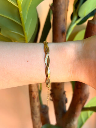 A close-up of a person's forearm wearing a thin twisted gold bracelet, set against a background of green leaves and brown branches.