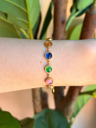 A gold-plated bracelet with five colorful gemstone beads (yellow, blue, green, pink, and orange) worn on a person's wrist, set against a background of green leaves.