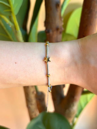 A delicate gold chain bracelet with a star charm and small gold beads, shown worn on a person's wrist against a background of green leaves and brown branches.