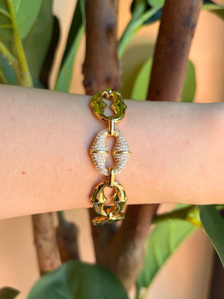 A gold-tone chain link bracelet with textured oval links and a central pavé diamond link, shown worn on a person's wrist against a background of green leaves and tree branches.