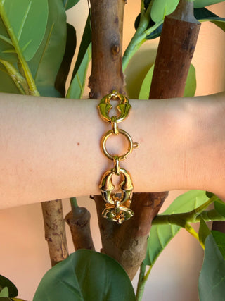 A gold-toned lobster claw charm bracelet with a key ring and a small bell, shown worn on a person's wrist against a background of green leaves and tree branches.
