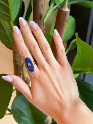 A close-up of a hand with pale pink manicured nails wearing a gold ring with a blue oval stone featuring a small yellow diamond in the center, set against a background of green leaves and brown branches.