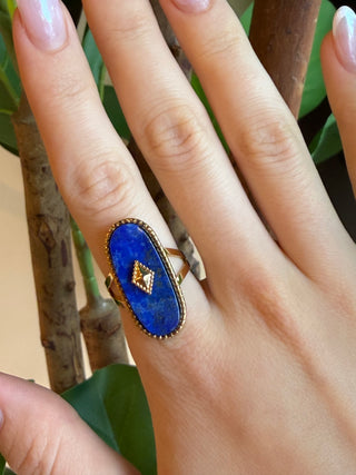 A gold ring with an oval blue lapis lazuli stone and a small central gold pyramid detail, shown worn on a person's finger.