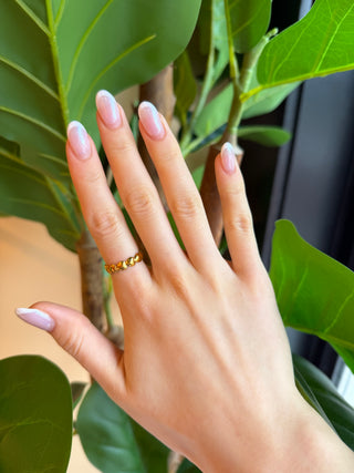 A close-up of a hand with manicured nails wearing a thin gold ring with a textured, raised pattern, set against a background of green plant leaves.