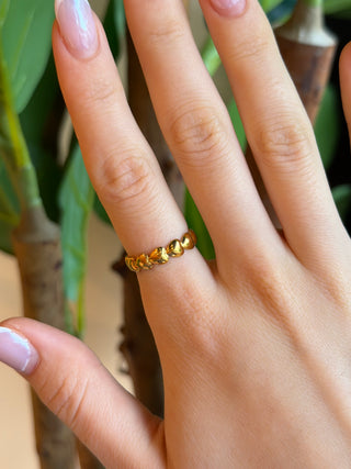 A close-up of a hand wearing a delicate gold ring with a textured, ridged band, set against a soft-focus background of green leaves and brown branches.