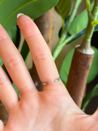 A close-up of the back of a gold adjustable ring with green leaves and brown branches blurred in the background.