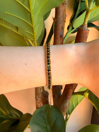 A gold chain bracelet with dark green faceted beads worn on a person's wrist, set against a background of green leaves and warm sunlight.