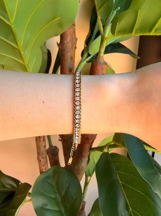 A gold tennis bracelet with clear stones worn on a person's wrist, set against a background of green leaves and warm sunlight.