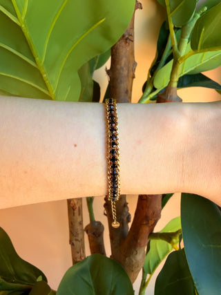 A delicate gold chain bracelet with black beads worn on a person's wrist, set against a background of green leaves and tree branches.