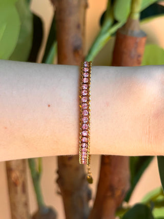 A delicate gold chain bracelet with small pink gemstones, worn on a person's wrist against a background of green leaves and brown tree trunks.