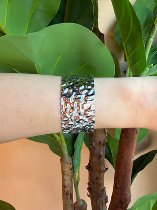 A silver-toned textured cuff bracelet worn on a person's wrist, shown against a background of green leaves and brown tree trunks.